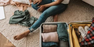 Personne assise sur un tapis en train de faire sa valise avant un voyage, avec vêtements soigneusement pliés, baskets blanches et accessoires prêts à être emportés.