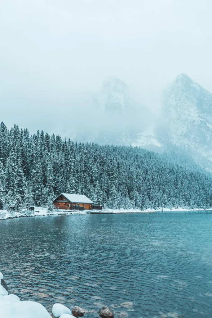 Chalet en bois au bord d’un lac gelé entouré de sapins enneigés, lieu idyllique à recommander dans un travel planner Canada pour une escapade hivernale en pleine nature.