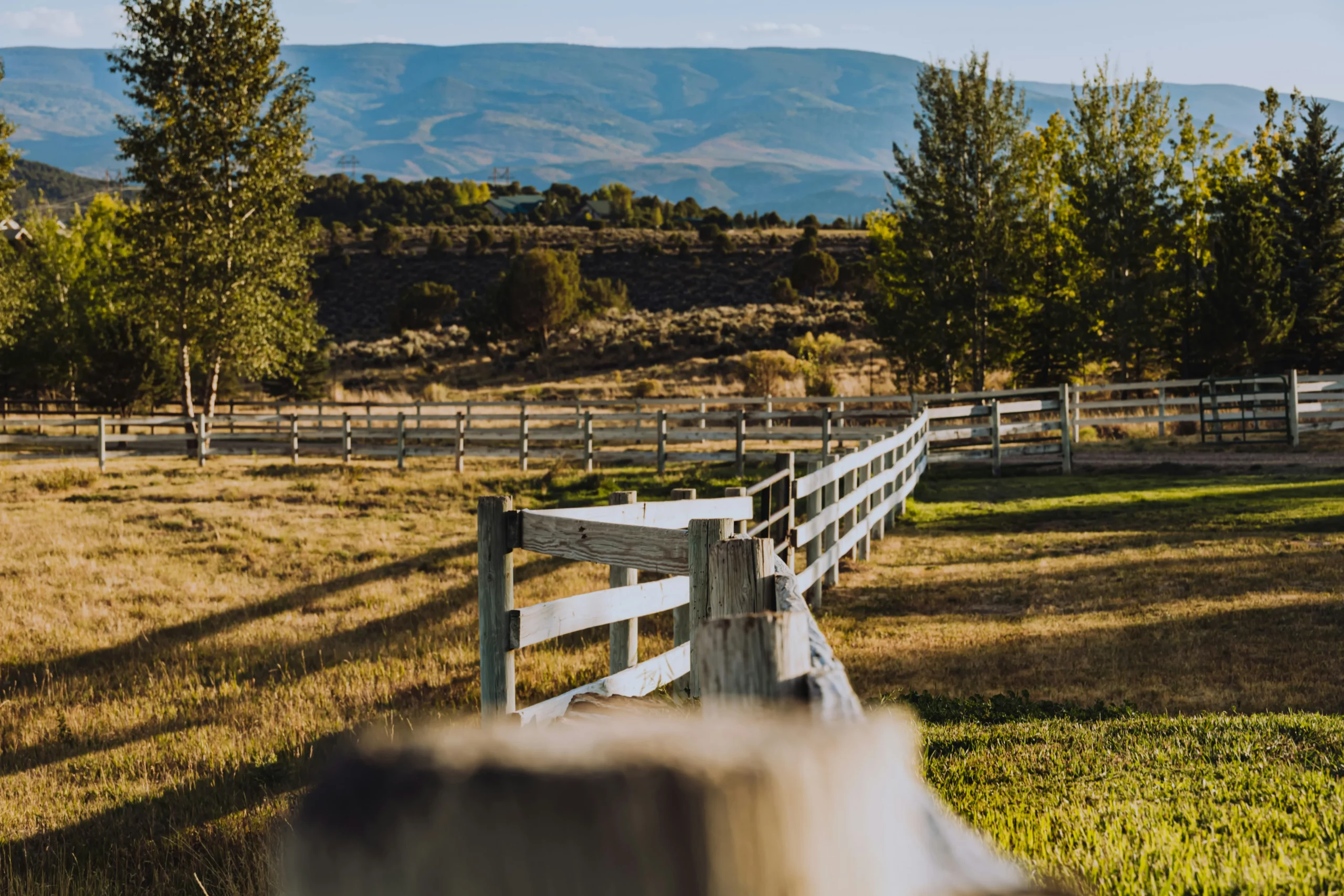 Clôture en bois devant un paysage rural avec ranch et montagnes à l’arrière-plan, évoquant la vie typique de l’Ouest américain pour un travel planner authentique.